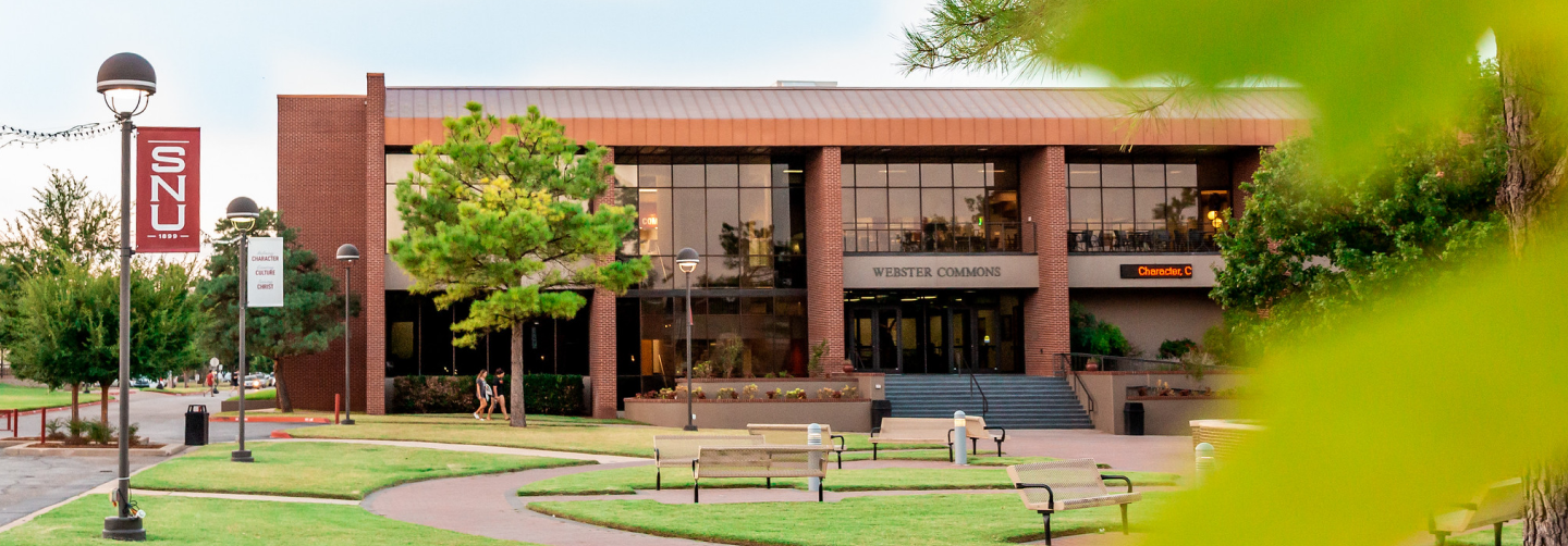 Southern Nazarene University Webster Commons building with bright green trees and benches in the foreground, showcasing a welcoming campus atmosphere.